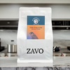 A white coffee bag with two description cards, the first one is multicoloured with a Falcon on it and the second says Strength Medium for the coffee card, the black logo for ZAVO is visible at the bottom of the bag. This coffee bag is on a marble work surface in a kitchen, with kitchen pans and utensils in the background.