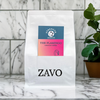 A white coffee bag with a black ZAVO logo stamped at the bottom of the bag, two product cards are visible for the Blend which is the Flamingo and the tasting notes which says Strength Medium, the image is shot on a marble backdrop with a plant in the background.