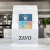 A white coffee bag with a black ZAVO logo stamped at the bottom of the bag, two product cards are visible for the blend which is the Hawk and the tasting notes, the image is shot on a marble kitchen work surface and in the background you can see a blurred kitchen and a plant