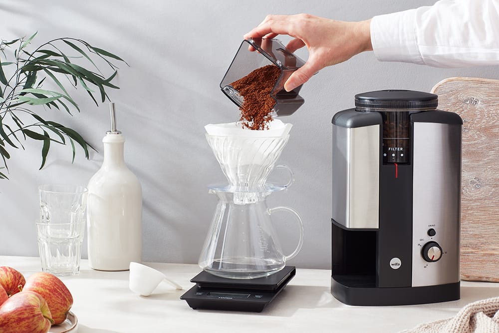 Ground coffee going in to a filter paper to make a cup of coffee, shot in a kitchen with glasses and apples in the background. The silver Wilfa Svart Coffee Grinder is visible.