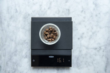 A black Felicita Incline scale on a marble background. The scale has beans in it showing a measurement of  16.1