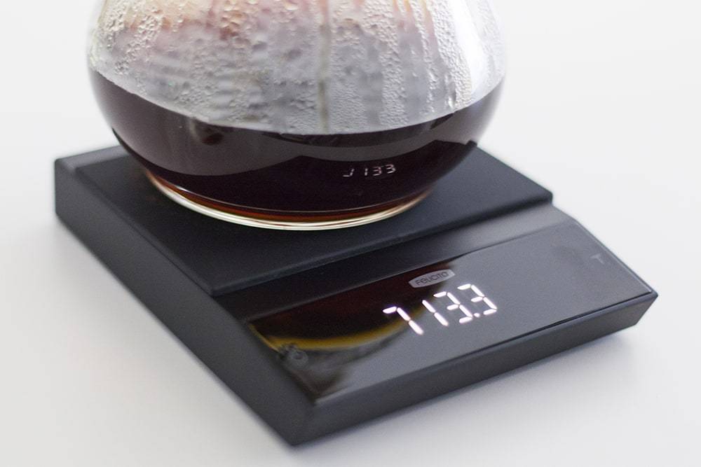 A black Felicita Incline scale with a glass pot of coffee on the top, the scale shows the value of 713.3